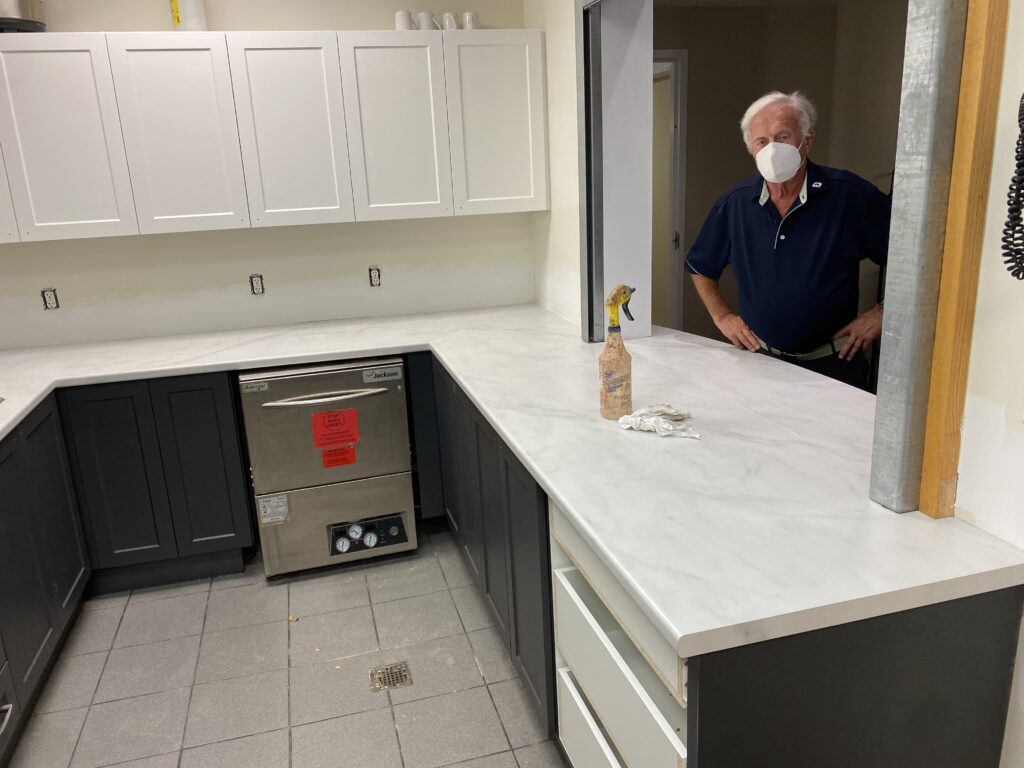 A man wearing a mask stands in a kitchen with white countertops and cabinets.